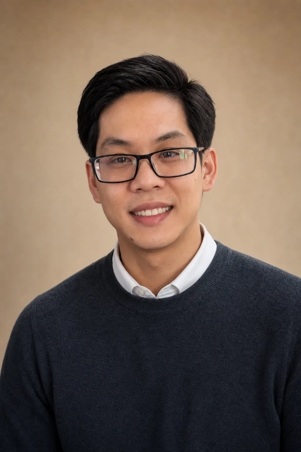 A man wearing glasses, a white collared shirt, and a dark sweater is smiling slightly in front of a plain beige background.